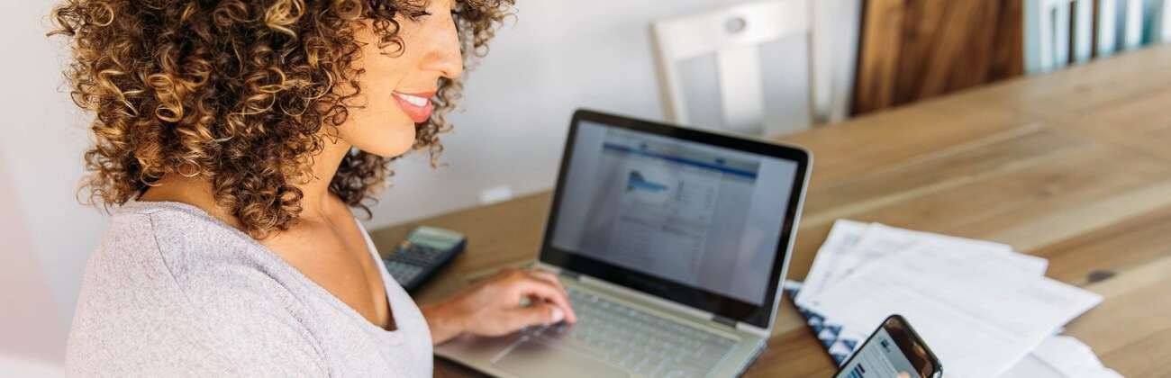 Woman works on bills with her laptop while glancing at her mobile device